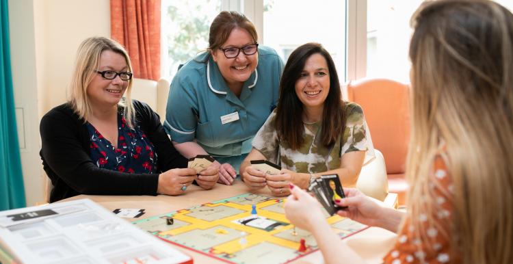 Three woman and a nurse sitting round a desk in a hospital day room playing a board game