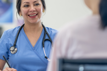 A woman in blue scrubs and wearing a stethoscope smiling at another person.